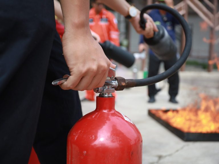 Hand hält einen Feuerlöscher, während ein Feuer im Hintergrund brennt.